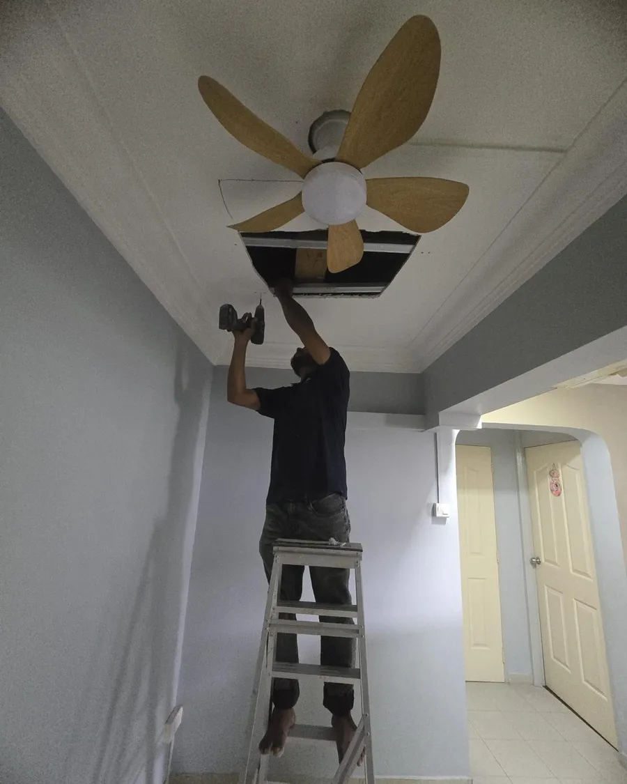 Technician on a ladder fixing a wooden-blade ceiling fan to the ceiling of an HDB corridor in Singapore
