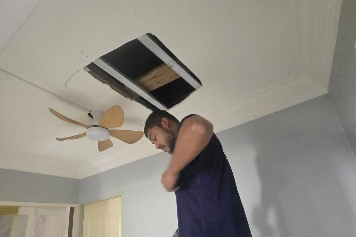 Technician working at an open ceiling hatch beside a newly installed wooden-blade ceiling fan in Singapore